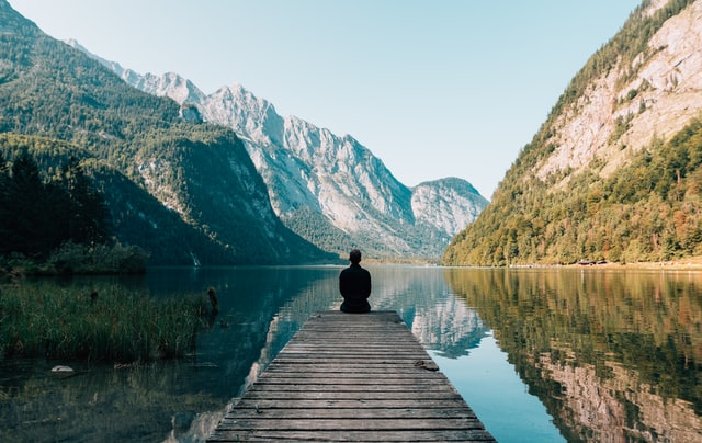 Man sitting on a dock overlooking the water with mountains in the background.