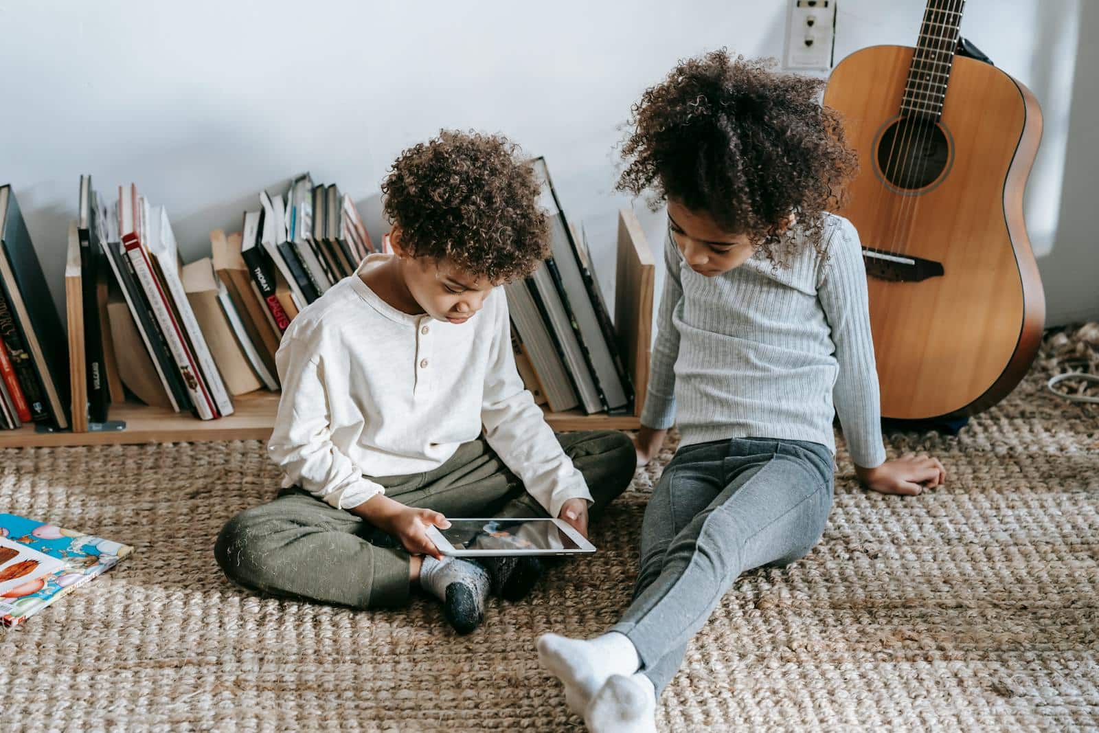 Thoughtful African American children in casual clothes sitting on floor against guitar and books and using tablet at home in daytime