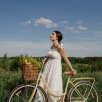 Woman in white dress standing with bicycle and basket in lush green meadow under blue sky.