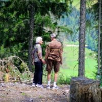 An elderly couple holding hands strolls through a serene forest trail, enjoying nature.