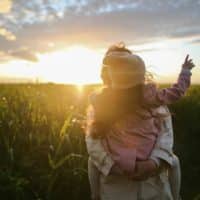 A mother and daughter embrace and point at the sunset in a grassy field.