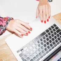 Close-up of woman's hands typing on a laptop with vibrant red nails, showcasing colorful floral sleeves.