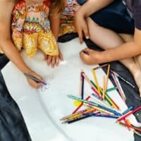 Two kids drawing on a picnic blanket with colored pencils outdoors.