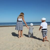 Three children in striped clothing walking on a sandy beach under a clear blue sky.