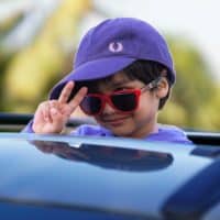 A cheerful child wearing a purple cap and red sunglasses makes a peace sign from a car rooftop.
