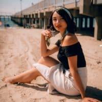 Chic woman in summer attire sitting on a sandy beach with a pier backdrop, exuding elegance.