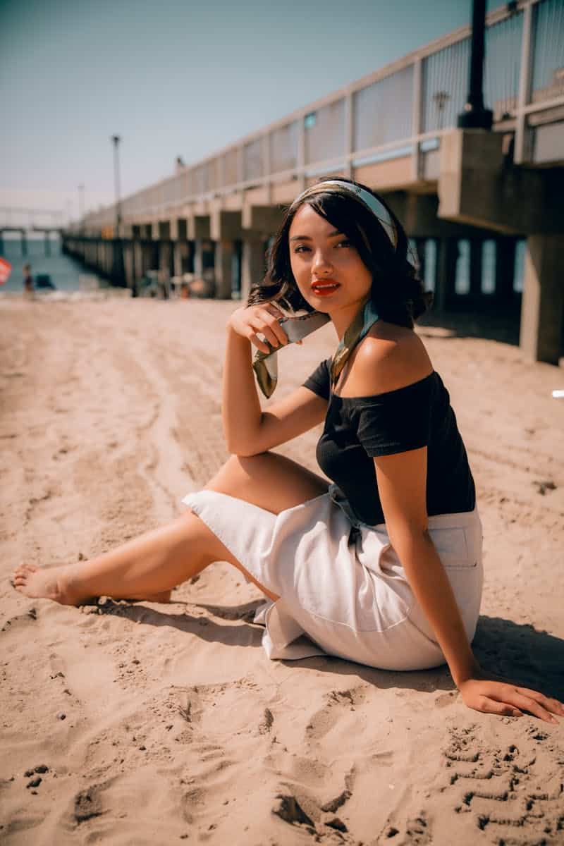 Chic woman in summer attire sitting on a sandy beach with a pier backdrop, exuding elegance.