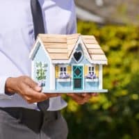 Businessman holds small house model outdoors, symbolizing real estate investment.