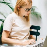 Focused young woman typing on a laptop in a bright, indoor setting with plants and papers.