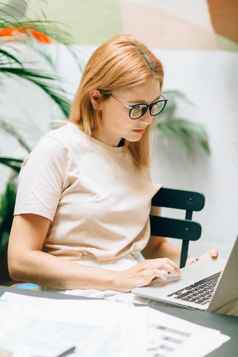 Focused young woman typing on a laptop in a bright, indoor setting with plants and papers.