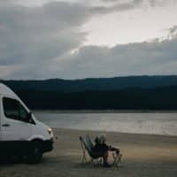 A woman relaxes by a lakeside, seated next to a campervan with a dog and folding chair under cloudy skies.