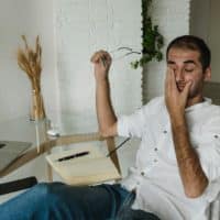 A tired man in a white shirt rests at a home office desk, holding eyeglasses and rubbing his eyes.