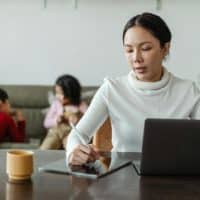 Focused young Asian woman in casual wear writing with stylus on tablet and browsing laptop while working remotely at home with children