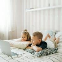 Two kids attentively watching a laptop on a bed, creating a cozy indoor setting.