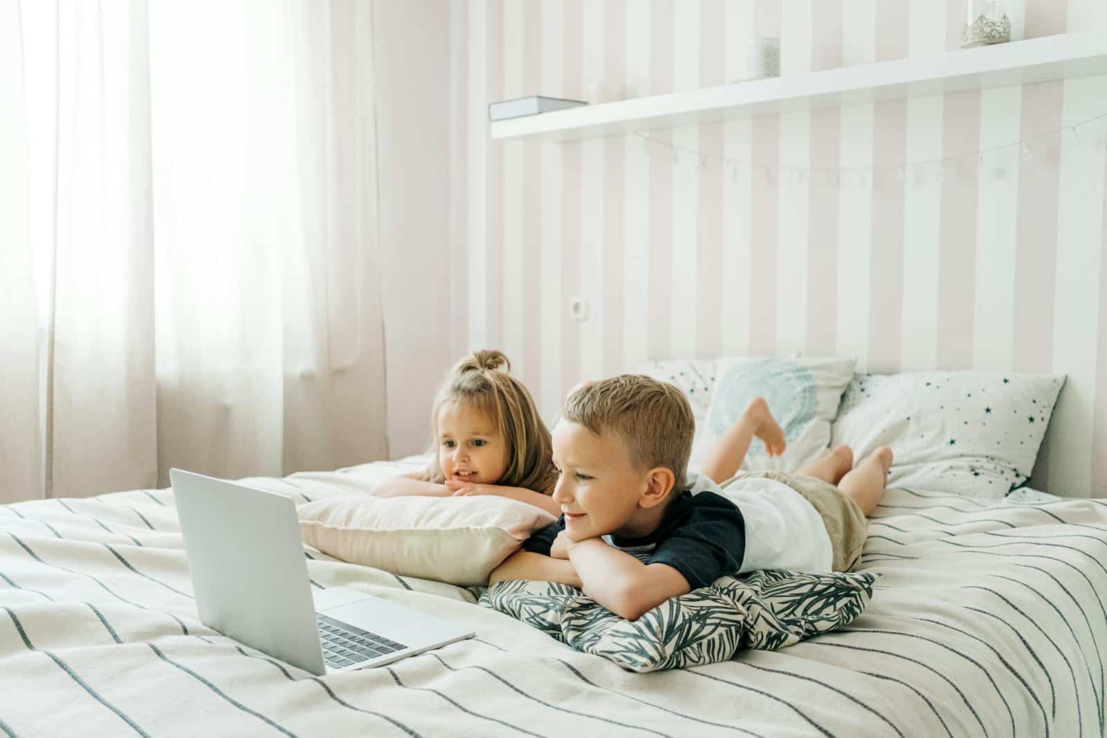 Two kids attentively watching a laptop on a bed, creating a cozy indoor setting.